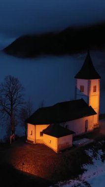 St. Primoz Kilisesi ve Felicijan Bulutlu Kış Gecesi 'nde aydınlatılmış. Bulutlu Gökyüzü. Jamnik, Slovenya, Avrupa. Hava görüntüsü. İnsansız hava aracı yukarı ve geri uçuyor. Dikey Video