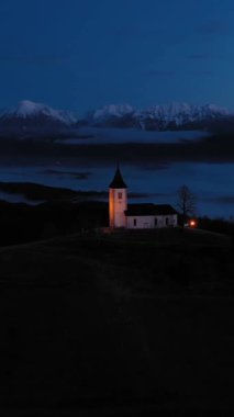 St. Primoz Kilisesi ve Felicijan Sabah Alacakaranlığı 'nda bulutların üstünde. Julian Alps. Jamnik, Slovenya, Avrupa. Hava görüntüsü. Yörüngede. Dikey Video