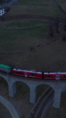 Akşamları İsviçre 'de Spiral Viaduct' ta tren. Bernina Demiryolu. İsviçre Alpleri. Hava görüntüsü. İnsansız hava aracı yukarı ve geri uçuyor. Dikey Video