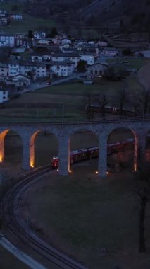 İsviçre 'deki Aydınlatılmış Spiral Viaduct Treni. Bernina Demiryolu. İsviçre Alpleri. Hava görüntüsü. İnsansız hava aracı yukarı ve geri uçuyor. Dikey Video