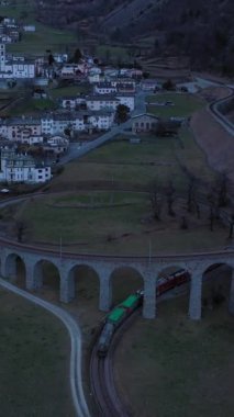 Akşamları İsviçre 'de Spiral Viaduct' ta tren. Bernina Demiryolu. İsviçre Alpleri. Hava görüntüsü. İHA ileri uçar, aşağı eğilir. Dikey Video