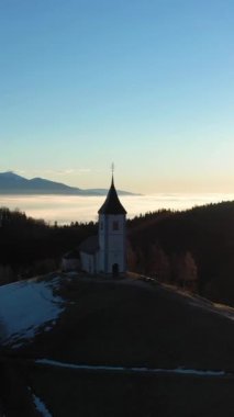 St. Primoz Kilisesi ve Felicijan Bulutların Üzerinde Gündoğumunda. Julian Alps. Jamnik, Slovenya, Avrupa. Hava görüntüsü. Yörüngede. Dikey Video