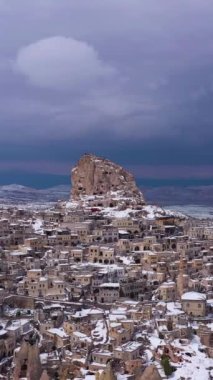 Taşa oyulmuş Uchisar Kalesi ve kışın Uchisar Şehri. Hindi. Karlı Tepeler. Hava görüntüsü. Fırtınalı Gökyüzü. Drone yörüngede. Dikey Video