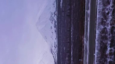 Kar yağışı sırasındaki Bulutlu Kış Günü 'nde Parklar Otobanı' nda ve Karlı Dağlar 'da araba. Alaska manzarası, ABD. Hava görüntüsü. İnsansız hava aracı yandan uçuyor. Dikey Video