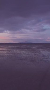 Sunset 'teki Uyuni Tuz Düzlükleri. Hava görüntüsü. Altiplano, Bolivya. Yağmur mevsimi. Tunupa Volkanı. Göl yüzeyinde Bulutların Yansıması. İHA alçaktan geriye doğru uçuyor. Dikey Video