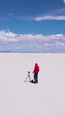 Tripod ve kameralı fotoğrafçı, güneşli bir günde Uyuni Tuz Düzlüklerine bakıyor. Hava görüntüsü. Altiplano, Bolivya. Kurak Mevsim. Altıgen Tuz oluşumları. Yörüngede. Solo Seyahat. Dikey Video