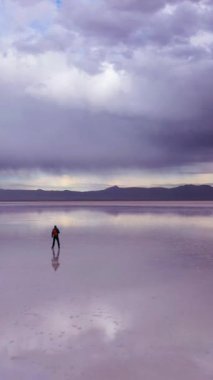 Adam ayakta duruyor ve Uyuni Tuz Düzlükleri 'ne bakıyor. Hava görüntüsü. Altiplano, Bolivya. Yağmur mevsimi. Suyun yansıması. Geniş açı. Dramatik ve Fırtına Bulutları. Yalnız Seyahat Konsepti. Dikey Video