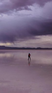 Turist ayakta duruyor ve Uyuni Tuz Düzlüklerine bakıyor. Hava görüntüsü. Altiplano, Bolivya. Yağmur mevsimi. Suyun yansıması. Yörüngede. Geniş açı. Dramatik Bulutlar. Yalnız Seyahat Konsepti. Dikey Video
