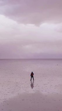 Yalnız turist ayakta duruyor ve Uyuni Tuz Düzlüklerine bakıyor. Hava görüntüsü. Altiplano, Bolivya. Yağmur mevsimi. Suyun yansıması. Yörüngede. Geniş açı. Dramatik Bulutlar. Solo seyahat. Dikey Video