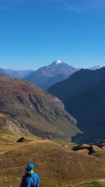 Mavi Ceketli Yürüyüşçü ayakta ve Dağlara bakıyor. Tepeler ve Dağlar. Albay Croix du Bonhomme. Fransız Alpleri, Fransa. Hava görüntüsü. Drone İleri Uçuyor. Dikey Video