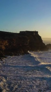 North Beach, Cliffs, Fort and Atlantic Ocean Sunset 'te. Nazare, Portekiz. Büyük Dalgalar. Hava görüntüsü. Drone geriye doğru uçuyor. Dikey Video