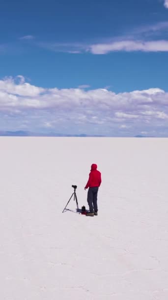 带着三脚架和照相机的摄影师在阳光明媚的日子里看着乌云盐滩 空中景观 玻利维亚阿尔蒂普拉诺 六角形盐层形成 独来独往垂直录像 — 图库视频影像