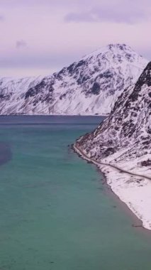 Yol, kışın Flagstadpolen ve Dağlar. Bulutlu Gökyüzü. Flakstadoya, Lofoten Adaları, Norveç. Hava görüntüsü. İnsansız Hava Aracı Yana Uçuyor, Eğil. Dikey Video