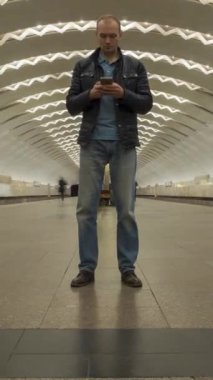Yer altındaki metro istasyonunda duran ve cep telefonu kullanan bir adamın zaman atlaması. Kalabalık insan ve geçen trenler. Dikey Video