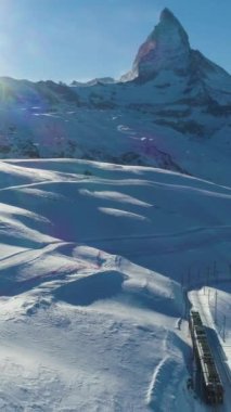 Matterhorn Dağı, Güneşli Kış Gününde Demiryolu ve Tren. İsviçre Alpleri. İsviçre. Hava görüntüsü. Şutu göster. İHA geriye uçuyor, Kamera yukarı kalkıyor. Dikey Video