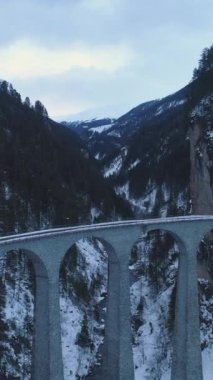 Kış Bayramı 'nda demiryolu ile Landwasser Viaduct. Mountain Gorge ve River. Hava görüntüsü. İsviçre Alpleri, İsviçre. Drone ileri, yukarı, kamera aşağı doğru uçuyor. Dikey Video