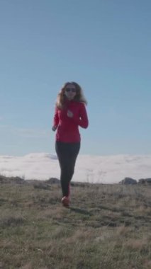 Güneş gözlüklü, uzun saçlı beyaz bir kadın bulutların üstünde koşuyor. Dikey Video