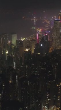 Gece vakti Hong Kong Skyline ve Victoria Harbour 'u aydınlattı. Hava görüntüsü. İHA ileri uçuyor, Kamera yukarı eğiliyor. Atış ayarlanıyor. 60 fp. Dikey Video