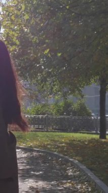 Gri paltolu, beyaz atkılı ve kıvırcık saçlı, güneyli güzel esmer kadın kameraya doğru yürüyor ve parkta dönüyor. Ağır çekim. Dikey Video
