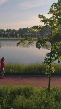 Sağlıklı genç bir kadın güneşli bir sabahta gölette güzel bir parkta koşuyor. Havadan görüş. Ağır çekim. Dikey Video