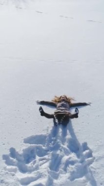 Genç ve neşeli kadın kara düşüyor ve kar melekleri yapıyor. Ağır çekim. Hava görüntüsü, ileri doğru uçuyor. Dikey Video