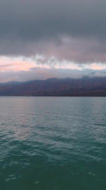 Gün batımında alçak açı gölün üzerinden sahile doğru uçuyor. Tepeler arka planda. Bulutlu, karanlık, fırtınalı bir gökyüzü ve güneş ışığı. Tekne manzarası. Dikey Video