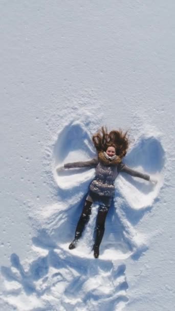 若い陽気な女性は雪の上に横たわり 雪の天使を作っています スローモーション 回転する空中ビュー バーティカルビデオ — ストック動画