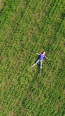 Kadın kocaman yeşil çimlerde uzanıyor ve dinleniyor. Drone dönüyor ve yukarı doğru uçuyor. Hava görüntüsü. Dikey Video