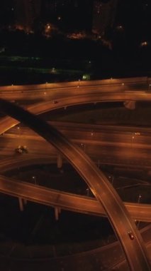 Aydınlatılmış karmaşık yol kavşağı ve gece araba trafiği. Drone geri dönüyor. Hava görüntüsü. Dikey Video
