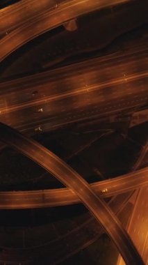 Aydınlanmış çok katlı yol kavşağı ve gece araba trafiği. Drone dönüyor ve uçuyor. Havadan dikey çekim. Dikey Video