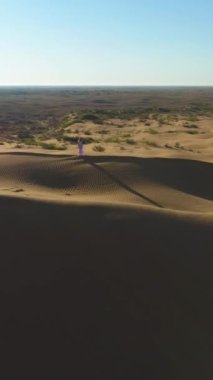 Drone, çölde kumlu bej kumulun tepesinde uzun saçlı, uzun saçlı, yalnız bir kadının üzerinde uçuyor. Hava görüntüsü. Dikey Video