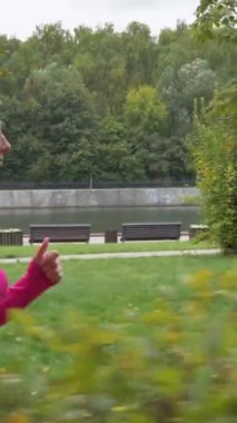 Genç Kafkasyalı kadın çalılar boyunca yeşil parkta koşuyor. Ağır çekim. Kamera hareket ediyor. Sabit çekim. Yan görüş. Dikey Video
