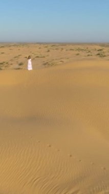 Beyaz elbiseli, uzun saçlı yalnız bir kadın çöldeki kum bej kum tepeciğinde kalıyor. Hava görüntüsü. Dikey Video