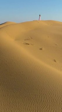 Beyaz elbiseli, uzun saçlı yalnız bir kadın çöldeki kum bej kum tepesinde kalıyor. Drone yaklaşıyor. Hava görüntüsü. Dikey Video