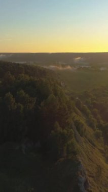Tepenin yamacında, güneş doğarken ağaçlar ve nehirle birlikte. Drone ileri uçuyor. Hava görüntüsü. Dikey Video