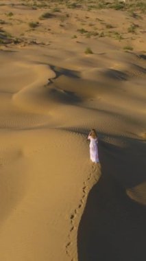 Beyaz elbiseli, uzun saçlı yalnız bir kadın çölde kumlu bej bir kum tepesinde yürüyor. Hava görüntüsü. Kamera aşağı eğiliyor. Dikey Video
