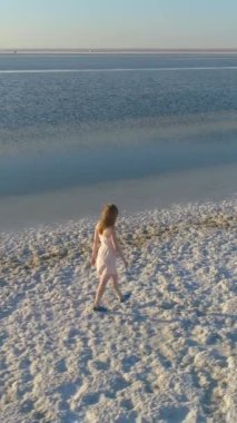 Beyaz elbiseli genç yalnız kadın tuz gölünün kıyısında yürüyor. Hava görüntüsü. Dikey Video