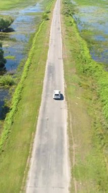 Beyaz araba bataklıktaki bir yola doğru gidiyor. Drone takip modunda. Hava görüntüsü. Dikey Video