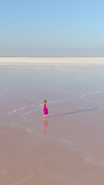 長い髪の赤いドレスの若い白人女性だけが巨大な塩湖で水の上を歩いています ドローンが前方に飛んでいる 空からの眺め バーティカルビデオ — ストック動画