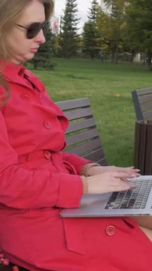 Güneş gözlüklü ve kırmızı pelerinli beyaz bir kadın şehir parkındaki bankta dizüstü bilgisayarla çalışıyor. Ön planda kırmızı çiçekler. Steadicam hareketli atış. 60 fp. Dikey Video