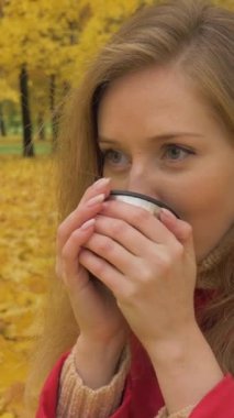 Kırmızı ceketli beyaz kadın termos metal bardaktan sıcak içecek içiyor. Arka planda sonbahar sarı yapraklar ve ağaçlar var. Orta boy. Dikey Video