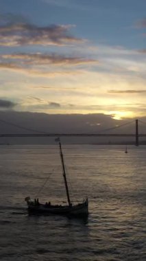 Sunset 'teki 25 Nisan Askılık Köprüsü ve oradan geçen yat silueti. Ponte 25 de Abril ve Tagus Nehri. Portekiz. Hava Sabit Çekimi. Altın Saat. Dikey Video