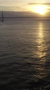 Sunset 'te 25 Nisan Askı Köprüsü. Ponte 25 de Abril ve Tagus Nehri. Portekiz. Hava görüntüsü. Altın Saat. İnsansız hava aracı ileri, yana doğru hareket ediyor. Şutu göster. Dikey Video