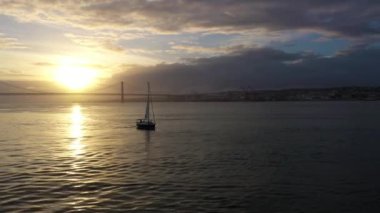 Sunset 'te 25 Nisan Askı Köprüsü ve Yelkenli Yat' ın Silueti. Ponte 25 de Abril ve Tagus Nehri. Portekiz. Hava görüntüsü. Altın Saat. Yörüngede