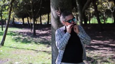Güneş gözlüklü at kuyruklu adam mızıkayla müzik çalıyor, Mouth Organ. Güneşli yaz gününde Green Park 'ta dikilmek. Orta Çekim