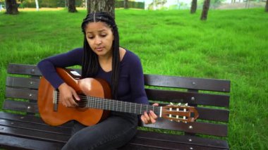 Kahverengi derili Asyalı Afrikalı Genç Kadın ve Yeşil Park 'ta Bench' te oturup gitar çalan uzun siyah saçlı. Tam Vuruş