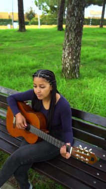 Kahverengi derili Asyalı Afrikalı Genç Kadın ve Yeşil Park 'ta Bench' te oturup gitar çalan uzun siyah saçlı. Tam doz. Dikey Video