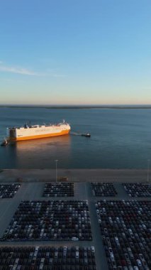 Ro-Ro Kargo Gemisi Römorkörle geliyor ve Port 'taki Sunset' te Büyük Tam Otopark Parkı 'nda yeni arabaların sıraları var. Otomotiv endüstrisi. Setubal, Portekiz. Hava görüntüsü. Yanlamasına hareket ediyor. Dikey Video