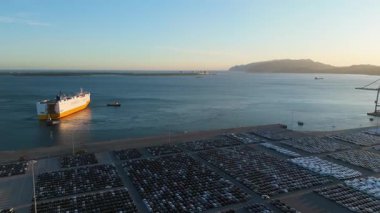 Ro-Ro Kargo Gemisi Römorkörle geliyor ve Port 'taki Sunset' te Büyük Tam Otopark Parkı 'nda yeni arabaların sıraları var. Otomotiv endüstrisi. Setubal, Portekiz. Hava görüntüsü. İleriye Taşınma