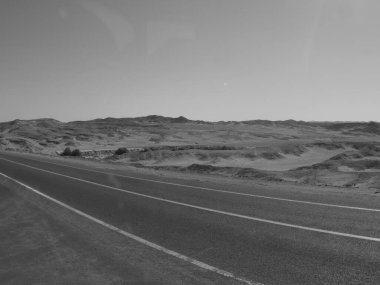 Çölde asfalt yol siyah ve beyaz renkte, dağların ortasında, gökyüzü bulutlarla ve siyah kumla kaplı.